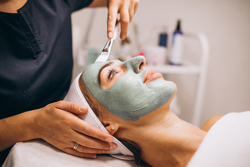 Spa esthetician gently applying facial mask to relaxed client on treatment table